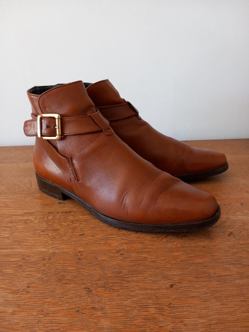 May include: A pair of brown leather ankle boots with a buckle closure and a gold-coloured buckle. The boots have a low heel and a rounded toe. The boots are displayed on a wooden surface.