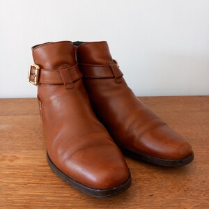 May include: A pair of brown leather ankle boots with a rounded toe and a small heel. Each boot features a strap with a gold-coloured buckle. The boots are set on a wooden surface.
