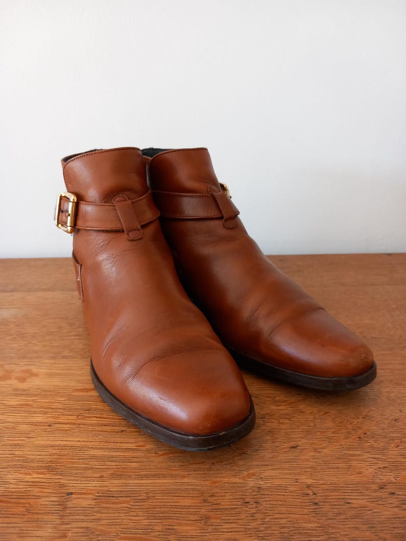 May include: A pair of brown leather ankle boots with a rounded toe and a small heel. Each boot features a strap with a gold-coloured buckle. The boots are set on a wooden surface.