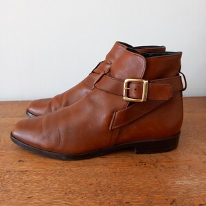 May include: A pair of brown leather ankle boots with a buckle closure and a gold-coloured buckle. The boots have a low heel and are set on a wooden surface. The boots are a classic style.