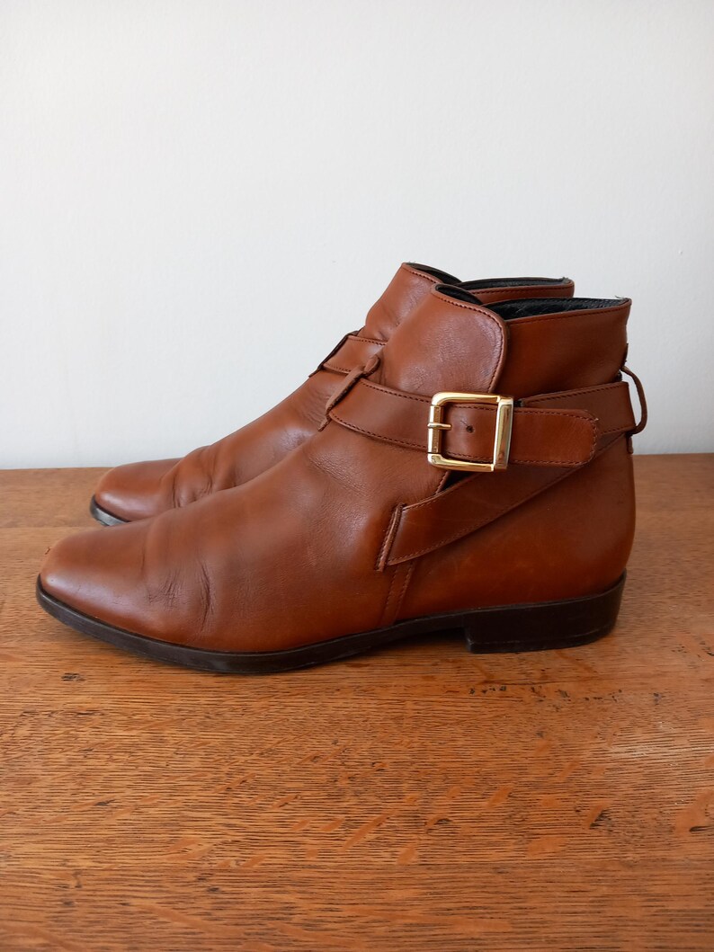 May include: A pair of brown leather ankle boots with a buckle closure and a gold-coloured buckle. The boots have a low heel and are set on a wooden surface. The boots are a classic style.
