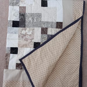 Może przedstawiać: Pikowana narzuta typu patchwork składająca się z różnych kwadratowych bloków materiału w odcieniach bieli, kremu, szarości i czerni z motywami kwiatowymi i geometrycznymi. Spód wykonany jest z beżowej tkaniny w ciemne kreski z granatową lamówką.