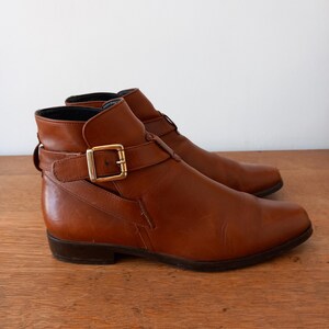 May include: A pair of brown leather ankle boots with a pointed toe and a gold-coloured buckle. The boots have a low heel and a strap that fastens around the ankle. The boots are displayed on a wooden surface.