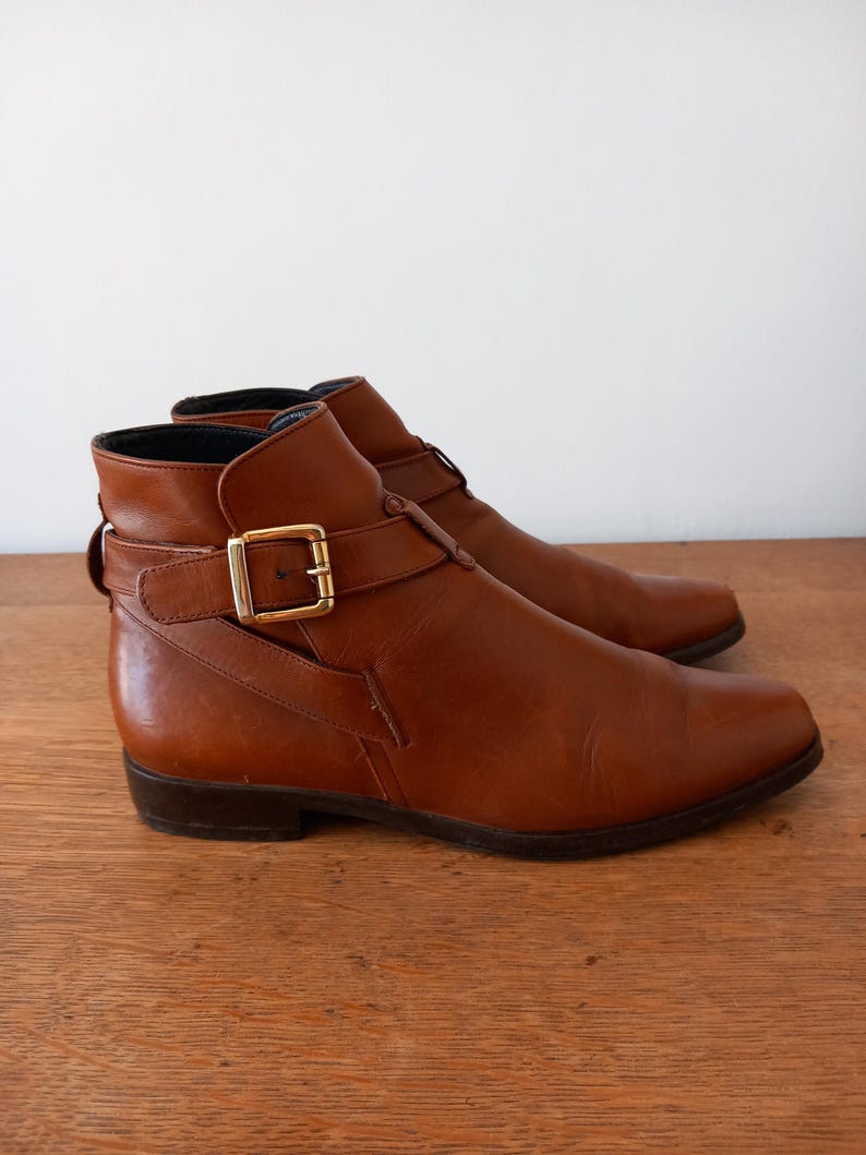 May include: A pair of brown leather ankle boots with a pointed toe and a gold-coloured buckle. The boots have a low heel and a strap that fastens around the ankle. The boots are displayed on a wooden surface.