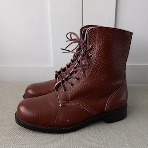 May include: A pair of brown leather lace-up boots featuring a textured grain finish, a rounded toe, and a black sole. The boots have dark brown laces threaded through metal eyelets, extending up the ankle with a high-top silhouette design.
