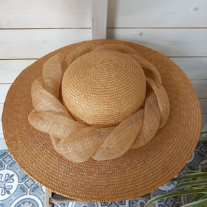 May include: A wide-brimmed sun hat in a natural straw color. The hat features a woven crown and a decorative band of woven material around the crown. The hat is displayed against a white wooden background.