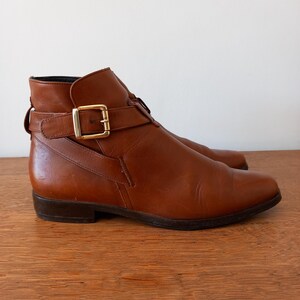 May include: A pair of brown leather ankle boots with a buckle fastening. The boots have a low heel and a gold-coloured buckle. The leather appears smooth and has a slight sheen. The boots are displayed on a wooden surface.