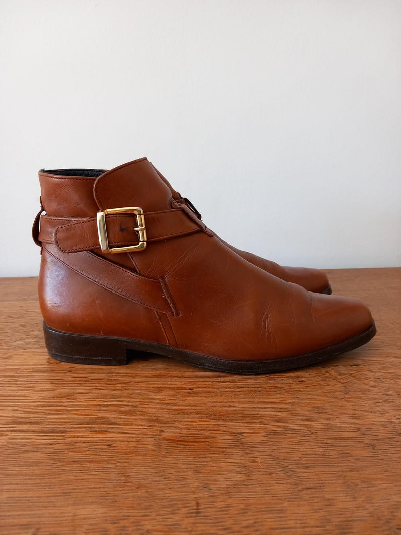 May include: A pair of brown leather ankle boots with a buckle fastening. The boots have a low heel and a gold-coloured buckle. The leather appears smooth and has a slight sheen. The boots are displayed on a wooden surface.