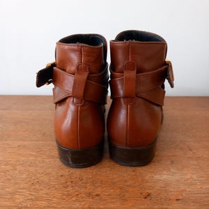 May include: A pair of brown leather ankle boots with decorative straps and gold-coloured buckles. The boots have a low heel and are set against a light background.
