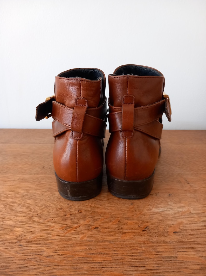 May include: A pair of brown leather ankle boots with decorative straps and gold-coloured buckles. The boots have a low heel and are set against a light background.