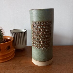 May include: A cylindrical ceramic vase with a matte sage green glaze and a textured, grid-like pattern of recessed squares and raised dots in a darker brown tone. The base of the vase is unglazed, revealing a light beige, natural clay finish.
