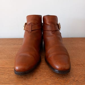 May include: A pair of brown leather ankle boots with buckle detailing. The boots have a rounded toe and a low heel. The leather has a slight sheen and the buckles are gold-coloured.