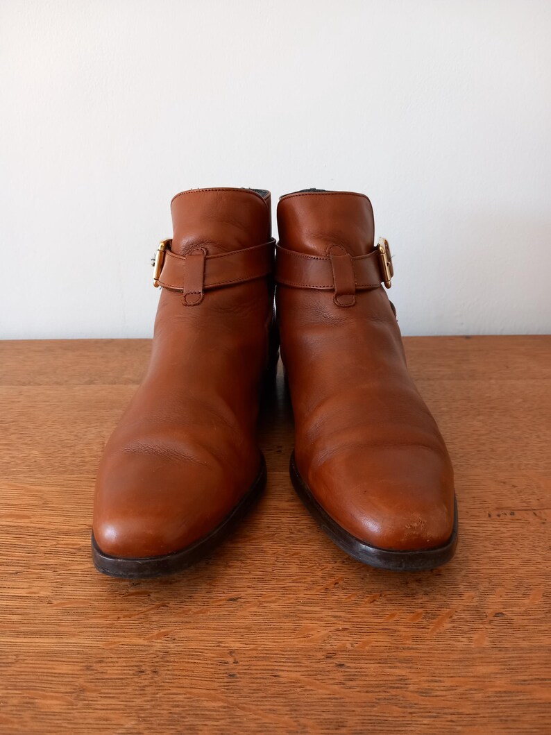 May include: A pair of brown leather ankle boots with buckle detailing. The boots have a rounded toe and a low heel. The leather has a slight sheen and the buckles are gold-coloured.
