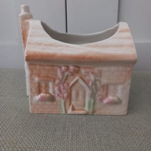 Op de afbeelding: Een huisvormige plantenbak van keramiek in een bleke perzik- en witte kleurstelling. De buitenkant heeft een gegoten baksteenstructuur, een centrale deur, twee ramen en verhoogde bloemendetails in zachte roze en groene tinten.