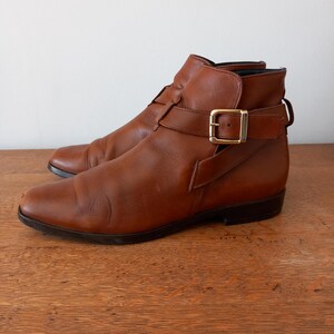 May include: Brown leather ankle boots with buckle fastening and a low heel. The boots feature a classic design with a pointed toe and an ankle strap. The leather has a worn appearance, giving a vintage feel. The boots are approximately 25 cm in length.