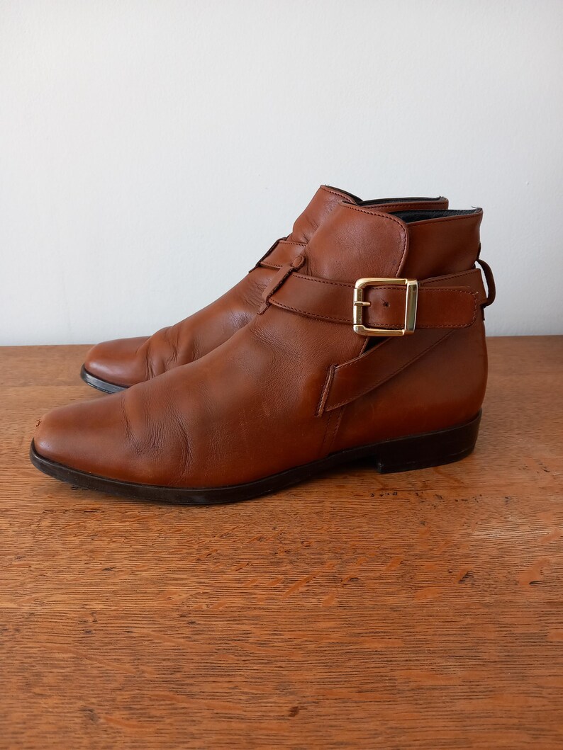May include: Brown leather ankle boots with buckle fastening and a low heel. The boots feature a classic design with a pointed toe and an ankle strap. The leather has a worn appearance, giving a vintage feel. The boots are approximately 25 cm in length.