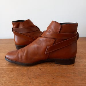 May include: A pair of brown leather ankle boots. The boots feature a pointed toe, a low heel, and a strap detail around the ankle. The leather has a slightly worn appearance.