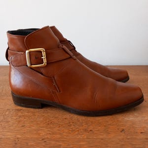 May include: Brown leather ankle boots with a buckle fastening. The boots feature a low heel and a strap detail around the ankle. The leather has a slight sheen, and the boots appear to be vintage.