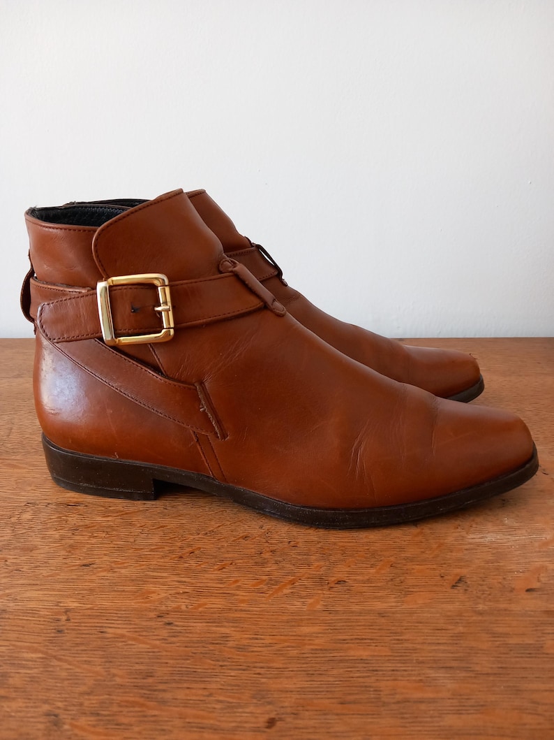 May include: Brown leather ankle boots with a buckle fastening. The boots feature a low heel and a strap detail around the ankle. The leather has a slight sheen, and the boots appear to be vintage.