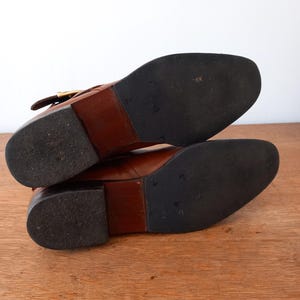 May include: A pair of brown leather boots with black rubber soles. The boots have a low heel and a buckle detail on the side. The soles show signs of wear.