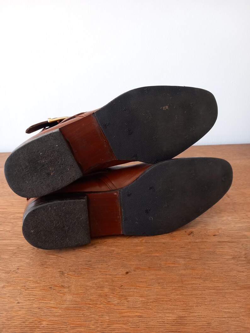 May include: A pair of brown leather boots with black rubber soles. The boots have a low heel and a buckle detail on the side. The soles show signs of wear.