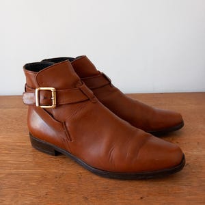 May include: Brown leather ankle boots with a pointed toe and a gold-coloured buckle. The boots have a low heel and a strap detail around the ankle. The leather has a smooth, polished finish.