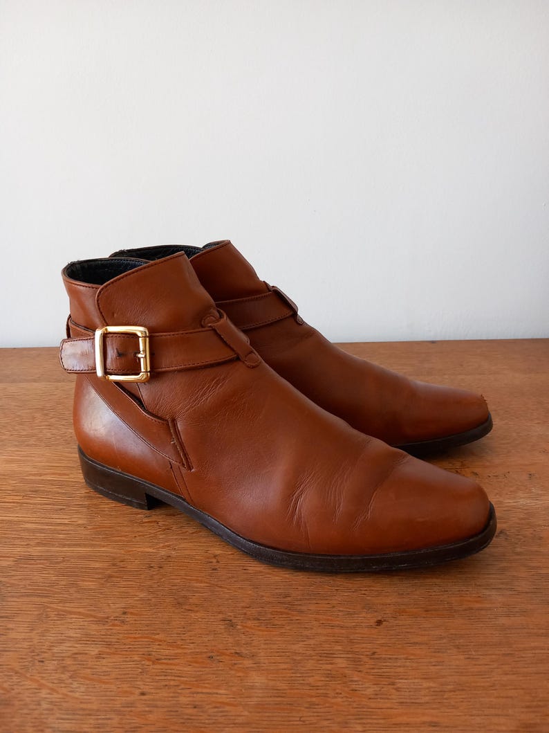 May include: Brown leather ankle boots with a pointed toe and a gold-coloured buckle. The boots have a low heel and a strap detail around the ankle. The leather has a smooth, polished finish.