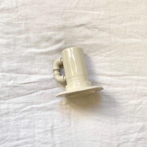 May include: A cream ceramic mug with an unusual handle. The mug has a cylindrical body and a wide, flat base. The handle is made of curved, pipe-like sections. The mug is set against a white, textured background.