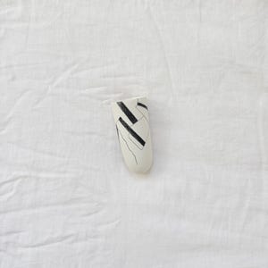 May include: A cylindrical ceramic wall vase with a matte off-white finish, featuring a hand-painted black graphic pattern of intersecting lines and geometric shapes on the front surface, with a flat top opening and a rounded bottom base.