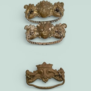 May include: Three antique brass drawer pulls with ornate designs. The top two pulls have a decorative face in the center and a curved handle. The bottom pull has a crown design. All three pulls are on a light blue background.
