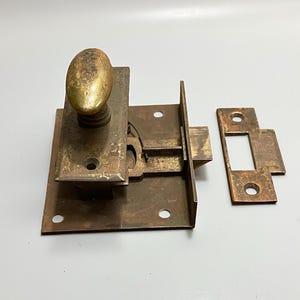 May include: An antique brass door latch set. The set includes a rectangular base with an oval knob, a latch mechanism, and a separate strike plate. The metal has a tarnished, aged appearance, suggesting vintage use. The set is designed for a door.