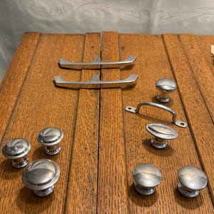 May include: A collection of silver-toned cabinet hardware, including knobs and handles, arranged on a wooden surface. The hardware features various designs, such as rounded knobs and curved handles, suitable for furniture restoration or new builds.
