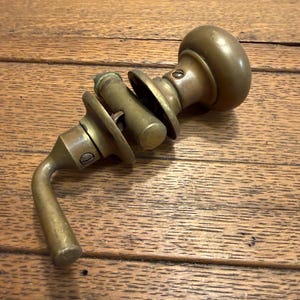 May include: An antique brass door handle set, featuring a round knob, a cylindrical spindle, and a lever handle. The set is tarnished, showing signs of age and use. The hardware is displayed on a wooden surface.