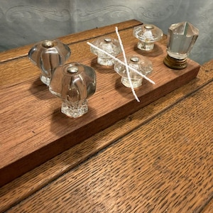 May include: A collection of five clear glass knobs with faceted surfaces and metallic hardware, arranged on a rectangular wooden board. Two thin white strings are crossed over the center knobs, and one knob features a gold-toned base.