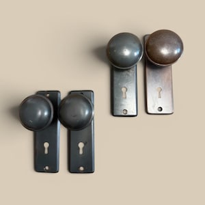 May include: Four vintage door knobs with keyhole plates. Two knobs are a dark gray color, and two are a bronze color. The knobs are round and the plates are rectangular. The door hardware is likely made of metal.