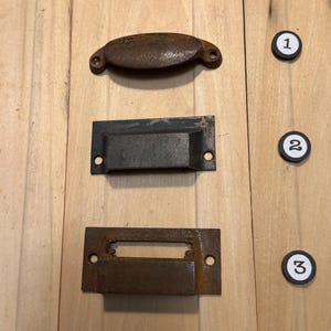 May include: Three antique metal drawer pulls in varying designs. The top pull is a curved handle with two screw holes. The middle pull is a rectangular recessed handle. The bottom pull is a rectangular pull with a central opening. All pulls have a rusty brown finish.