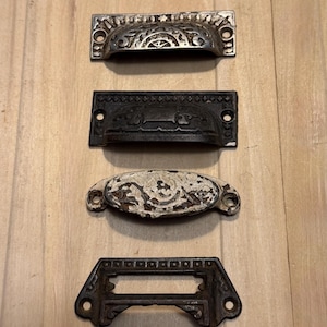 May include: Four antique drawer pulls in various styles. The top pull is silver-toned with ornate detailing. Below it is a black pull with a curved design. The third pull is oval-shaped with white paint. The bottom pull is a dark metal frame.