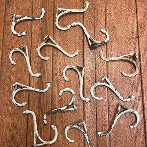 May include: A collection of antique white metal hooks with a distressed finish, arranged on a wooden surface. The hooks have a curved design and show signs of wear, suggesting they are vintage or reclaimed items.