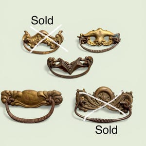 May include: Five ornate metal drawer pulls with swinging handles, arranged in a group. Two of the pulls are marked with a white X and the word Sold. The hardware features various decorative embossed patterns, aged brass tones, and curved designs.