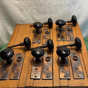 May include: A collection of antique door knobs and plates. The knobs are black and round, with a glossy finish. The plates are rectangular with keyholes and a tarnished, bronze-like appearance. The set is arranged on a wooden surface.