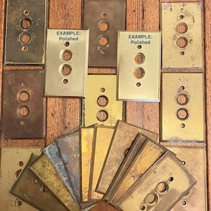 May include: A collection of vintage brass light switch plates in various shades of gold and brown. Some plates have three circular openings and are labeled "EXAMPLE: Polished". The plates show signs of age and wear, suggesting they are antique or reclaimed.