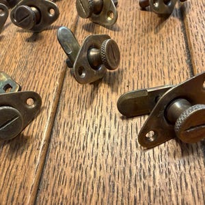 May include: Close-up of several antique brass latches with screw-in knobs and lever arms. The latches are set against a wooden surface, showcasing their aged patina and intricate design. The knobs have a slotted head.