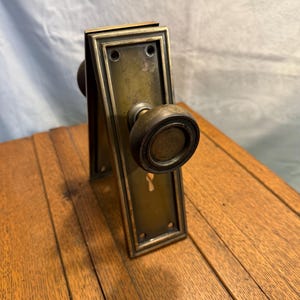 May include: Antique brass door knob set with a rectangular backplate and a round knob. The set has a keyhole and is mounted on a wooden surface. The brass has a vintage patina.