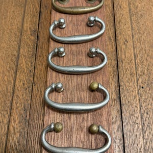 May include: Five antique drawer pulls in various finishes. The top pull is brass, while the others are silver-toned. Each pull has a curved handle with decorative end pieces. The pulls are arranged vertically on a wooden surface.
