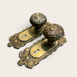 May include: Two antique brass door knobs with ornate detailing. Each knob has a round, textured design and is mounted on a rectangular backplate with a keyhole. The brass has a warm, aged patina, suggesting vintage charm.