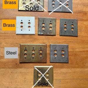 May include: A collection of rectangular metal wall plates for light switches, featuring various finishes including brass and steel. Some plates are marked with the word Sold and have white X marks over them. The plates have multiple circular cutouts.