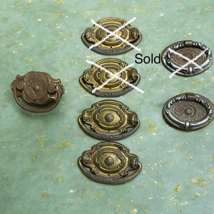 May include: A collection of seven metal drawer pulls. One ornate round pull is on the left. A vertical column of four oval brass pulls is in the center, with two marked with white X's. Two oval pulls with silver-toned rings are on the right.