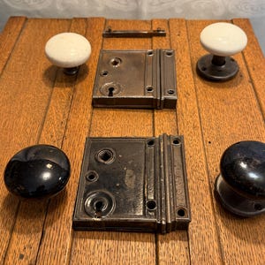 May include: Antique door hardware set featuring two white porcelain doorknobs, two black doorknobs, two metal door latches, and two metal strike plates. The hardware is arranged on a wooden surface.