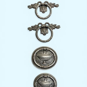 May include: Four antique silver-toned drawer pulls. The top two pulls have a ring design with leaf accents. The bottom two pulls are round with a detailed urn design. All pulls are arranged vertically against a light blue background.