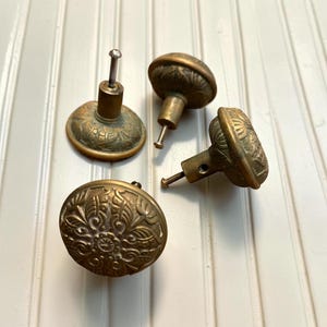 May include: Four round brass cabinet knobs featuring an intricate embossed floral pattern on the face. Each knob has a central cylindrical post and a threaded metal screw extending from the back for attachment to furniture or cabinetry doors.